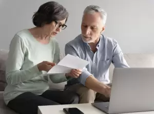 Pareja de adultos mayores sentados en el sofá revisando documentos y utilizando una computadora portátil, discutiendo posiblemente sobre la tributación seguros de vida y otros asuntos financieros de planificación de jubilación.