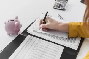 Vista de cerca de una persona escribiendo notas en un cuaderno, junto a un cerdito de alcancía y documentos de impuestos. La imagen simboliza la importancia de la planificación fiscal y la contabilidad al preparar la declaración de la renta.