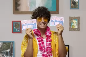 Joven sonriente con camisa de verano, gafas de sol amarillas y un collar de flores, sosteniendo varios boletos de "rasca y gana" de lotería. Esta lotería instantánea es diferente a la Lotería de Navidad, pero ambos premios están sujetos a Hacienda.