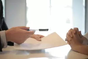 $$\text{Detalle de las manos de un asesor legal o gestor ofreciendo un bolígrafo para la firma de la documentación clave en el proceso de traspaso de negocio en España.