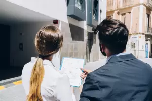 Una pareja analiza los planos de una casa mientras observa una vivienda. La asesoría de profesional previene problemas futuros de humedades y goteras al comprar una propiedad.
