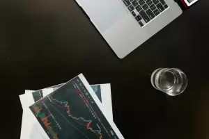 Overhead view of a laptop, financial charts, and a glass of water on a desk, illustrating the complexity of crypto tax analysis.