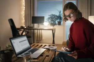A focused woman working remotely on a tablet and laptop in the evening, representing the dedicated work style of those considering Beckham Law for Remote Workers.